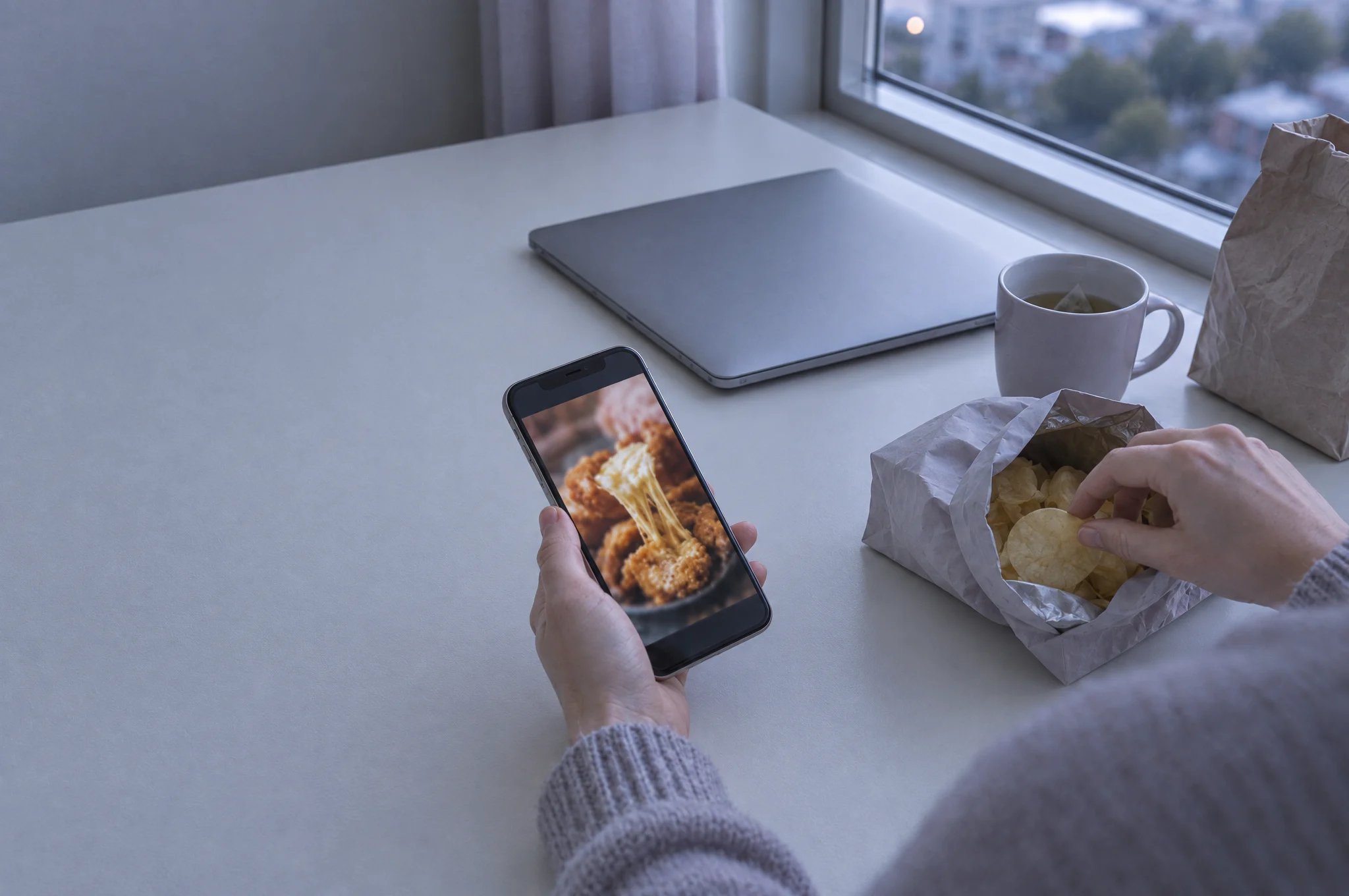 A desk by a window in the evening. One hand holds a phone playing a food reel, the other reaches into a packet of crisps. A closed laptop, a mug and a paper delivery bag sit nearby.